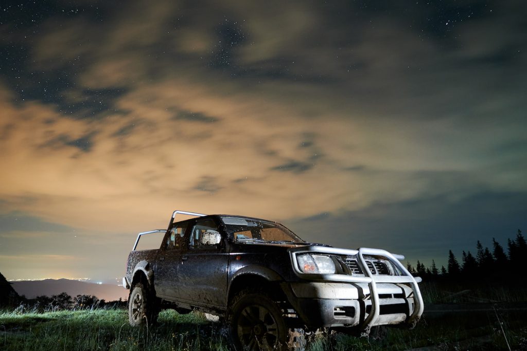 Illuminated-pickup-offroad-truck-in-mud-on-mountain-meadow-at-night-under-starry-cloudy-sky